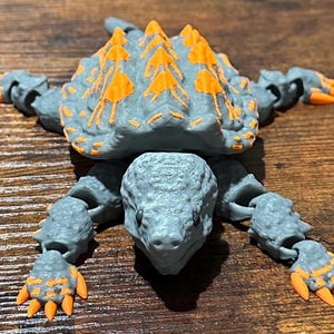 May include: A gray and orange articulated toy ankylosaurus dinosaur. The toy has a textured, armored back with orange accents. The dinosaur's limbs and head are also gray with orange detailing on the claws.