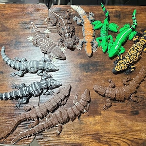 Peut inclure: Une collection de lézards jouets aux couleurs et motifs variés, dont vert, orange, noir, jaune, marron et gris. Les lézards sont disposés sur une surface en bois, mettant en valeur leurs motifs et textures détaillés.