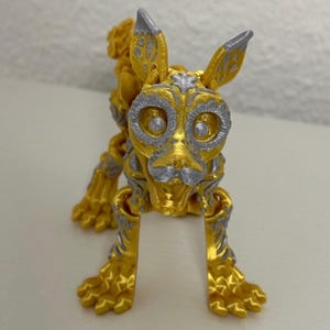 May include: A golden-yellow dog figurine with intricate silver-colored detailing. The dog has large eyes, pointed ears, and a detailed skeletal structure. The figurine is standing on all fours, showcasing its detailed paws.