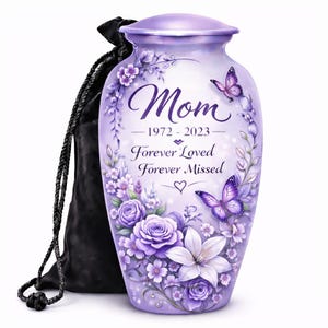 May include: A light purple cremation urn featuring a floral design with roses and lilies, two butterflies, and the text Mom 1972-2023 Forever Loved Forever Missed. The urn is accompanied by a black drawstring bag with a braided cord.