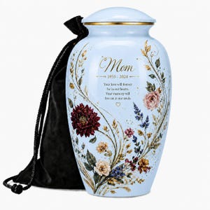 Könnte beinhalten: Hellblaue Urne mit goldenem Rand und Deckel, verziert mit floralen Motiven und dem Wort "Mom" mit den Daten 1955-2024. Eine schwarze Samttasche ist enthalten. Der Text lautet "Your love will forever be in our hearts. Your memory will live on in our souls."