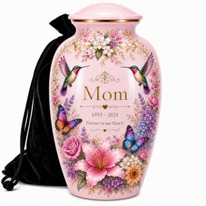 May include: A pink cremation urn with a floral design, hummingbirds, and butterflies. The urn has the word "Mom" and the years 1955-2024 printed on it. A black velvet bag is next to the urn.