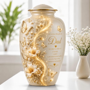 Könnte beinhalten: Cremefarbene Urne mit goldenem Deckel, verziert mit goldenen Schmetterlingen, Blumen und einem leuchtenden Rankenmuster. Der Text lautet "In Loving Memory of Dad" mit einer herzlichen Botschaft. Ein Gedenkstück.