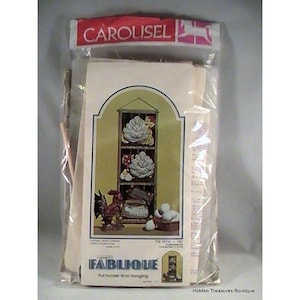 Könnte beinhalten: Eine versiegelte Verpackung mit einem Fabrique Topflappen-Wandbehang-Kit. Das Kunstwerk zeigt einen Hahn, Eier und dekorative Regale mit floralen Designs. Die Verpackung ist oben mit "CAROLISEL" beschriftet.