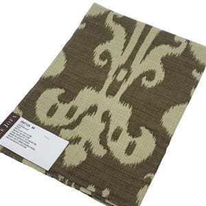 May include: A folded fabric sample with a repeating ikat pattern in shades of brown and beige. The fabric has a textured appearance and a tag with the text "LEE JOFA" and product information. The fabric is made in the United States.