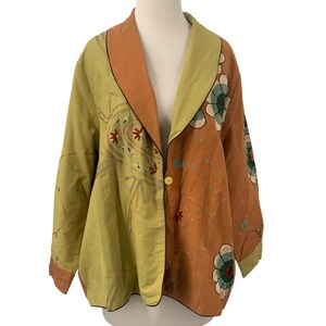 May include: A two-tone jacket with a unique design. The jacket features a combination of olive green and orange-brown colors. The olive green side has embroidered floral patterns, while the orange-brown side has floral and leaf patterns. It has a single gold button closure.