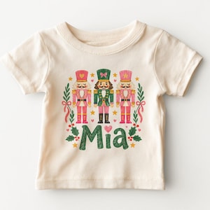 May include: A cream-colored short-sleeve t-shirt featuring a graphic of three nutcrackers in pink and green uniforms. The design includes holly leaves, small stars, and hearts, with the name Mia printed in large green textured letters below them.