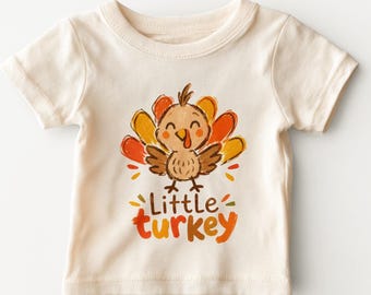 May include: A cream-colored short-sleeve t-shirt featuring a central graphic of a cartoon turkey with brown, orange, and yellow feathers. Below the turkey, the text Little turkey is printed in brown and orange lettering with small leaf accents.