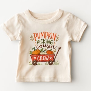 May include: A cream-colored short-sleeve t-shirt featuring a graphic of a red wagon filled with orange pumpkins. The text reads PUMPKIN PICKING COUSIN CREW in orange, green, and brown lettering, surrounded by small green leaves and grass.