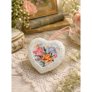 May include: A white heart-shaped trinket box with a floral design. The box features a pink cord for hanging and a detailed, embossed border. The center of the box displays a bouquet of colorful flowers, including pink, orange, and purple blooms.
