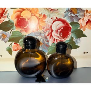 May include: Two round, amber-colored glass perfume bottles with black spherical caps. The bottles have a smooth, curved silhouette and contain a dark liquid. They are positioned side-by-side, with the larger bottle on the left and the smaller on the right.
