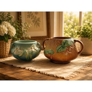 May include: Two ceramic vessels with handles, one smaller and light blue with white floral relief, the other larger and amber-brown with green leaf relief. Both feature rounded bodies and are positioned side by side on a textured fabric mat.