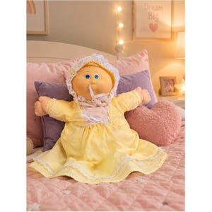 May include: A doll with a round face, blue eyes, and yarn hair, wearing a pale yellow dress with white lace trim and a matching bonnet. The doll has a soft, fabric body and is dressed in a light-colored outfit with a white ribbon tie at the neck.