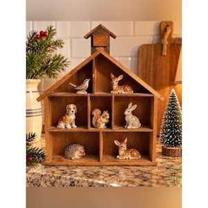 May include: A wooden house-shaped shelf with six compartments containing small ceramic animal figurines: a bird, a fawn, a dog, a squirrel, a rabbit, a hedgehog, and another fawn. The structure has a peaked roof and a brown wood finish.