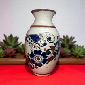 May include: Cream ceramic vase with a brown rim and base. The vase has a hand-painted design of a blue bird, blue flowers, and brown and green foliage. The vase has a bulbous body and a narrow neck.