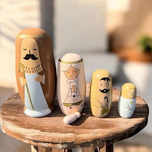 May include: A set of four wooden nesting dolls depicting a nativity scene. The dolls are painted in various colors, including brown, beige, and yellow, with detailed illustrations of religious figures. The dolls are arranged on a wooden surface.