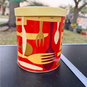 Puede incluir: Un recipiente de cocina cilíndrico vintage con tapa amarilla. El cuerpo rojo está decorado con diseños de utensilios dorados, blancos y burdeos. El recipiente es probablemente de metal y es un artículo de cocina retro.