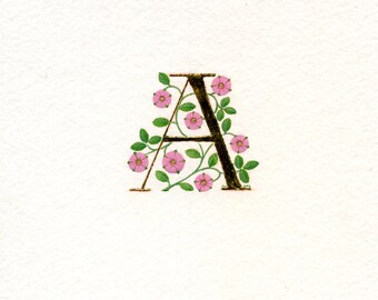 Puede incluir: Una letra A dorada con flores rosas y hojas verdes que la rodean.