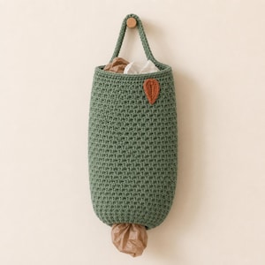 May include: A sage green crocheted hanging storage bag with a textured stitch pattern. The bag features a single top handle and a small orange leaf-shaped applique on the front. A brown plastic bag is visible protruding from the bottom opening.