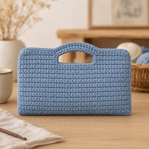 Peut inclure: Un sac à main en crochet bleu clair avec une poignée. Le sac rectangulaire est fait d'un matériau tricoté texturé. Le sac est posé sur une surface en bois.