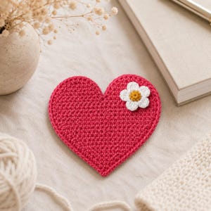 Peut inclure: Un dessous de verre au crochet en forme de cœur d'une couleur magenta vive. Le dessous de verre présente une petite application de fleur blanche à six pétales avec un centre jaune fixée sur le côté supérieur droit du cœur. La texture est dense.