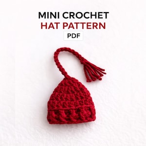 Könnte beinhalten: Ein kleiner, handgefertigter roter Häkelhut mit einer langen, geflochtenen Quaste. Der Hut ist eine Miniaturgröße mit einem strukturierten Stichmuster. Der Text auf dem Bild lautet "MINI CROCHET HAT PATTERN PDF".