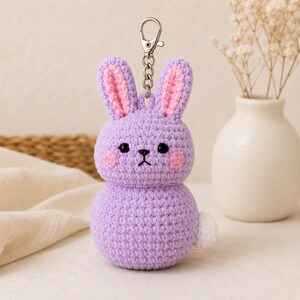 Peut inclure: Un porte-clés lapin en crochet avec un corps lavande, de longues oreilles aux accents roses, des yeux en perles noires, un nez brodé et des joues roses. Une petite queue blanche est visible sur le côté, avec un fermoir mousqueton en métal argenté.