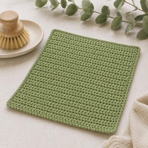May include: A square, sage green crocheted dishcloth featuring a dense, uniform textured stitch pattern. The cloth is positioned next to a round, light-colored ceramic dish holding a wooden scrub brush with light-colored natural bristles.