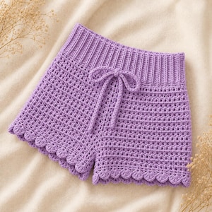 Peut inclure: Un short en crochet lavande avec une taille à cordon et des bords festonnés. Le short fait à la main présente un motif de point texturé et est présenté sur un fond neutre. Le short est de couleur violet clair.