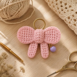 Peut inclure: Un porte-clés en forme de nœud papillon rose crocheté, avec un anneau doré et un petit charme de perle violette. Le nœud est fait de fil doux et a un aspect texturé. Le porte-clés est un accessoire mignon.