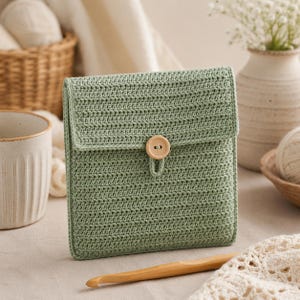 May include: A sage green crochet pouch with a flap closure and a wooden button. The pouch is rectangular and has a textured appearance. A wooden crochet hook is visible in the foreground.