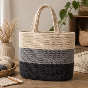 Peut inclure: Un sac cabas en fil crocheté présentant un design à blocs de couleurs avec trois bandes horizontales en crème, gris moyen et gris anthracite foncé. Le sac possède deux anses assorties en crochet de couleur crème et une base ovale structurée.