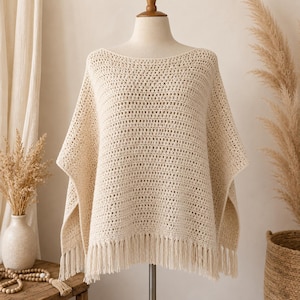 Pode incluir: Um poncho de crochê na cor creme com um decote canoa largo e um padrão de pontos abertos repetitivos. A peça possui uma bainha reta finalizada com franjas longas e pendentes que caem uniformemente ao longo da borda inferior do item.