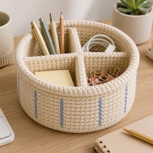 Peut inclure: Un organiseur de bureau rond en crochet de couleur crème, divisé en quatre compartiments. Il présente des rayures verticales bleu clair et contient des stylos, un crayon, des post-it, des trombones et un câble blanc. Un petit pot de plante succulente est visible en arrière-plan.
