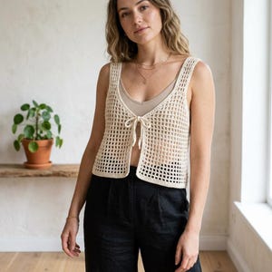 Peut inclure: Un gilet en crochet crème à tissage ouvert avec une fermeture à nouer. Le gilet est sans manches et porté sur un haut de couleur neutre. Le modèle porte un pantalon noir. Une petite plante dans un pot en terre cuite est visible en arrière-plan.