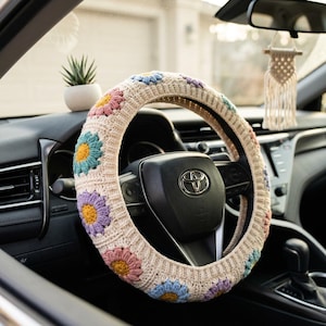 Peut inclure: Un couvre-volant en crochet beige orné de fleurs de marguerite colorées bleues, jaunes, roses et violettes. Le couvre-volant est installé sur un volant noir à l'intérieur d'une voiture. Une petite plante succulente en pot est sur le tableau de bord.