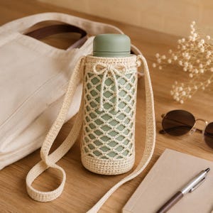 Peut inclure: Une gourde vert sauge dans un support en crochet crème avec une sangle assortie. Le support présente un motif en filet en forme de losange et un nœud en haut. La gourde est sur une surface en bois avec un sac fourre-tout, des lunettes de soleil et un carnet.