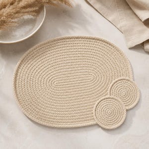 May include: A beige oval-shaped crocheted placemat accompanied by two matching circular crocheted coasters. The items feature a consistent, tightly stitched corded texture throughout, creating a uniform and structured appearance for table settings.