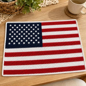 May include: A rectangular crochet placemat featuring the design of the United States flag. The piece displays a dark blue canton with white stars and alternating horizontal red and white stripes, all finished with a white crocheted border.