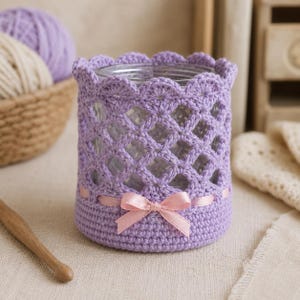 Peut inclure: Un bocal en verre transparent recouvert d'une housse en crochet lavande avec un bord festonné, un motif en losanges et une base pleine. Un fin ruban de satin rose clair est entrelacé dans le crochet et noué en un petit nœud.
