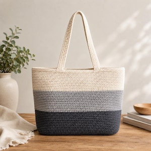 Peut inclure: Un sac fourre-tout en crochet de forme rectangulaire avec deux anses. Le sac présente trois blocs de couleurs horizontaux : crème en haut, gris au milieu et gris foncé en bas. Le sac est posé sur une surface en bois.