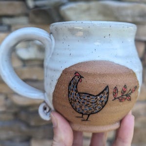 Puede incluir: Una taza de cerámica hecha a mano con una parte superior blanca moteada y una base marrón. La taza presenta una ilustración dibujada a mano de una gallina con detalles azules y negros y una cresta roja. Una ramita de hojas rojas y verdes está al lado de la gallina.