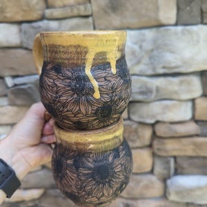 May include: Two handmade ceramic mugs stacked, held by a hand. The mugs feature a sunflower design etched in black on a brown base. The rims are glazed with a dripping yellow glaze. The mugs are approximately 4 inches tall.