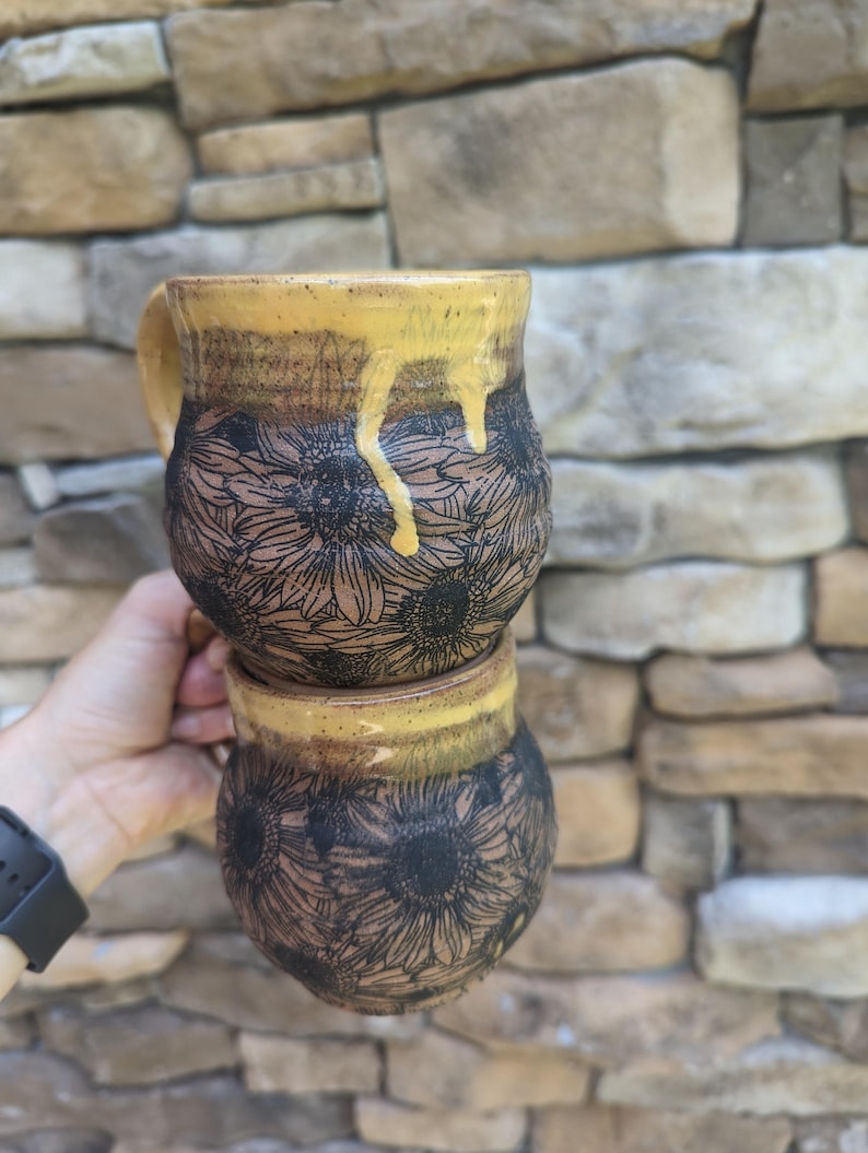 May include: Two handmade ceramic mugs stacked, held by a hand. The mugs feature a sunflower design etched in black on a brown base. The rims are glazed with a dripping yellow glaze. The mugs are approximately 4 inches tall.