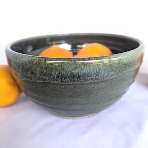 May include: A handmade, green ceramic bowl with a textured surface. The bowl is filled with bright orange fruit. The bowl has a wide, shallow shape and a dark green glaze with lighter green accents.