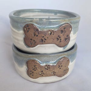 Puede incluir: Dos cuencos de cerámica para mascotas apilados uno encima del otro. Los cuencos son de color azul claro y blanco con un diseño en forma de hueso marrón. Los cuencos hechos a mano tienen un aspecto rústico. Cada cuenco mide unos 15 cm de diámetro.