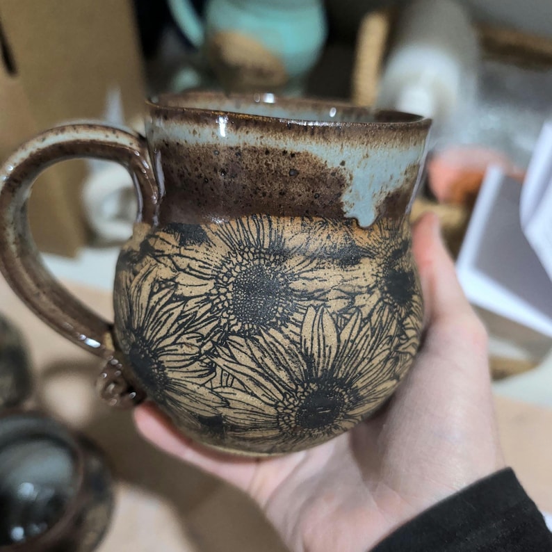 May include: A handmade ceramic mug with a brown handle and rim, featuring a sunflower design. The mug has a light brown base with black sunflower illustrations and a light blue interior rim. The mug is held in a hand.