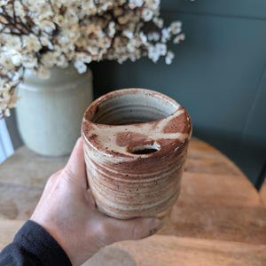 Puede incluir: Un portacepillos de cerámica hecho a mano con un diseño único. El soporte tiene forma cilíndrica con una parte superior curvada y un agujero para un cepillo de dientes. El color es una mezcla de beige y marrón, con un patrón texturizado y rayado.