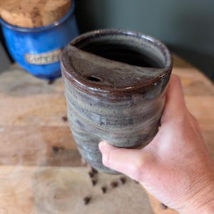 May include: A cylindrical ceramic travel mug with a speckled grey and blue glaze. The mug features a built-in ceramic lid with a small drinking aperture and a dark brown rim. The exterior surface has a textured, hand-thrown appearance with visible rings.