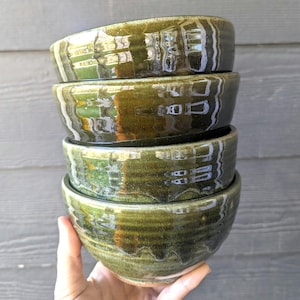 May include: A stack of four handmade ceramic bowls. The bowls are glazed in a glossy, olive green color with subtle variations. The bowls have a rounded shape and a textured surface, reflecting light.